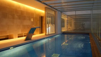 A swimming pool with tiled walls and a glass ceiling, surrounded by wooden deck and metal railings.