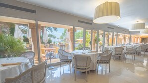 A spacious dining area with round tables and wicker chairs, large windows offering views of the outdoors, and a polished marble floor. - S'Agaró Hotel Spa & Wellness (Castell-Platja d'Aro)