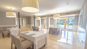 A spacious dining area with multiple tables and chairs, large pendant lights, and a view of the outdoor pool area. - S'Agaró Hotel Spa & Wellness (Castell-Platja d'Aro)
