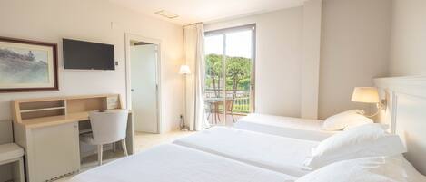 A hotel room with two beds, a desk, a chair, a TV, and a balcony with a view of greenery.