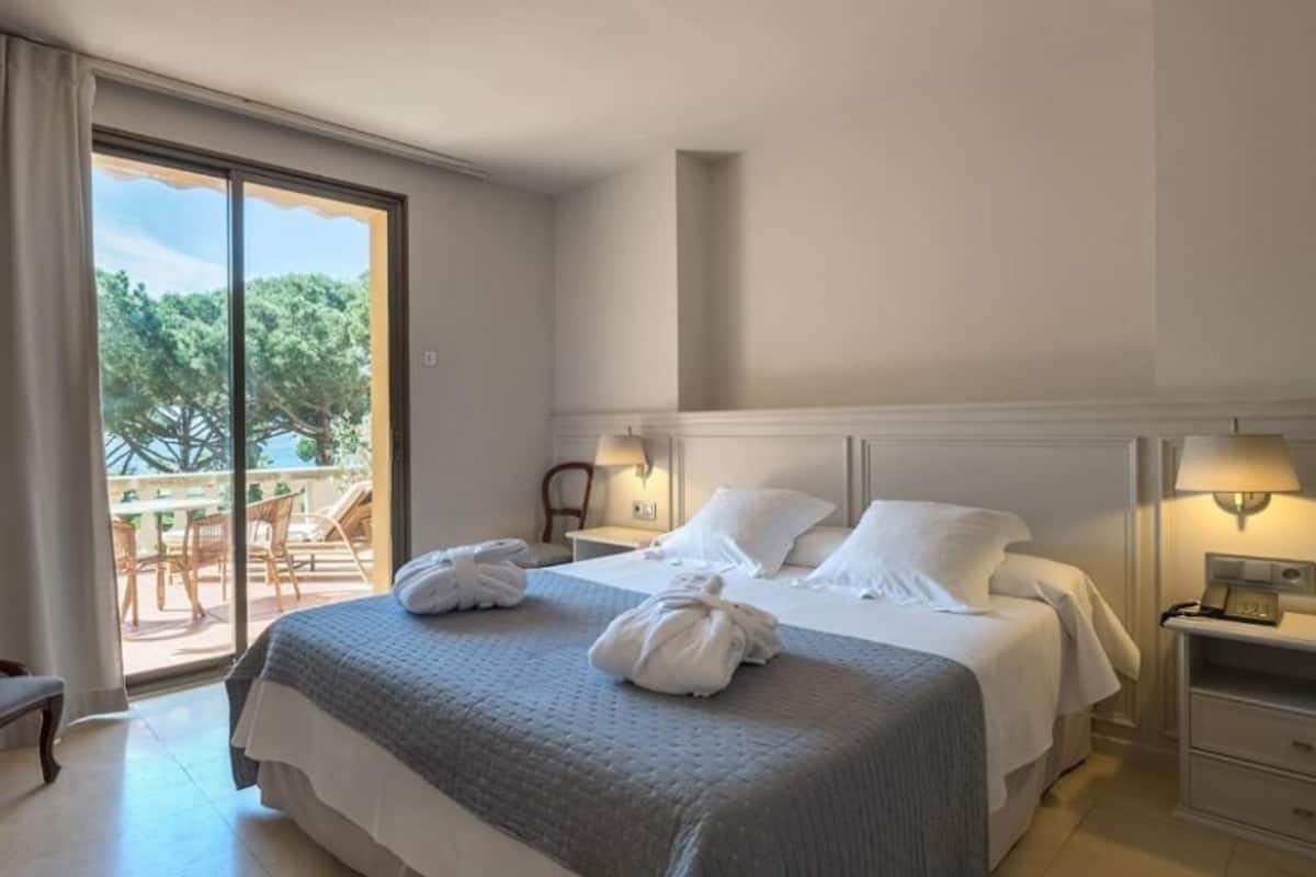 A hotel room with a large bed, two bedside tables with lamps, a chair, and a view of the outdoors through a sliding glass door.