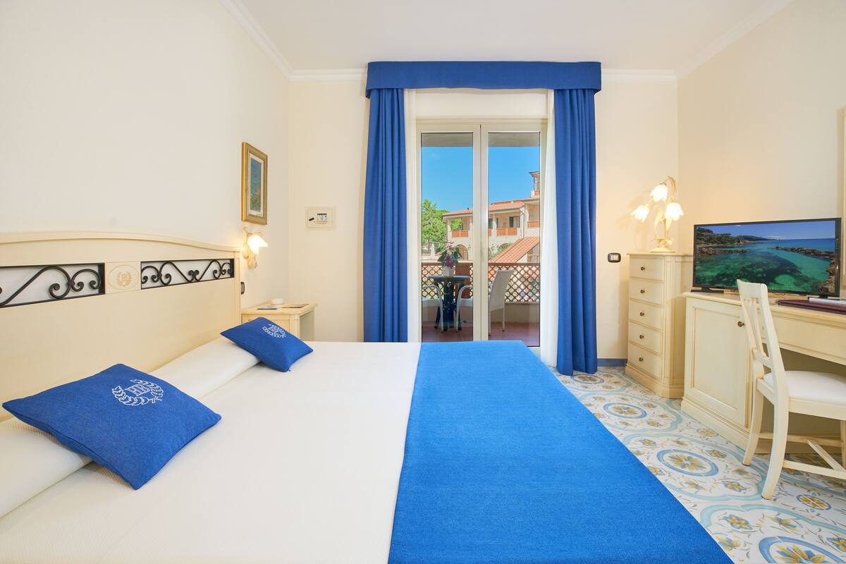 A hotel room with a bed, a desk, a chair, a TV, and a balcony with blue curtains.