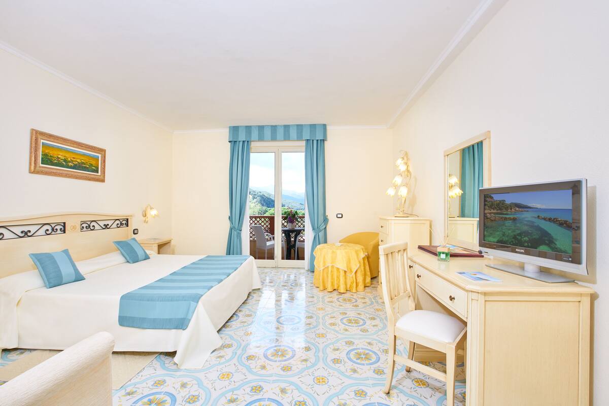 A hotel room with a bed, desk, chair, television, and a balcony with a view.