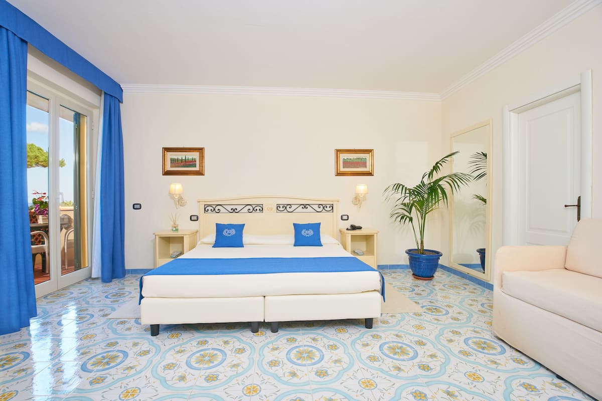A bedroom with a bed, a nightstand, a potted plant, a sofa, and a sliding glass door leading to an outdoor area.