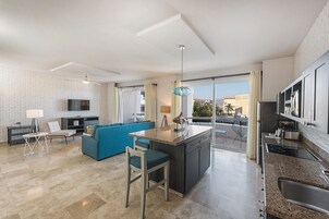 Una cocina moderna con una isla central, un rincón de comedor con un sofá azul y un amplio ventanal con vistas a edificios y palmeras.