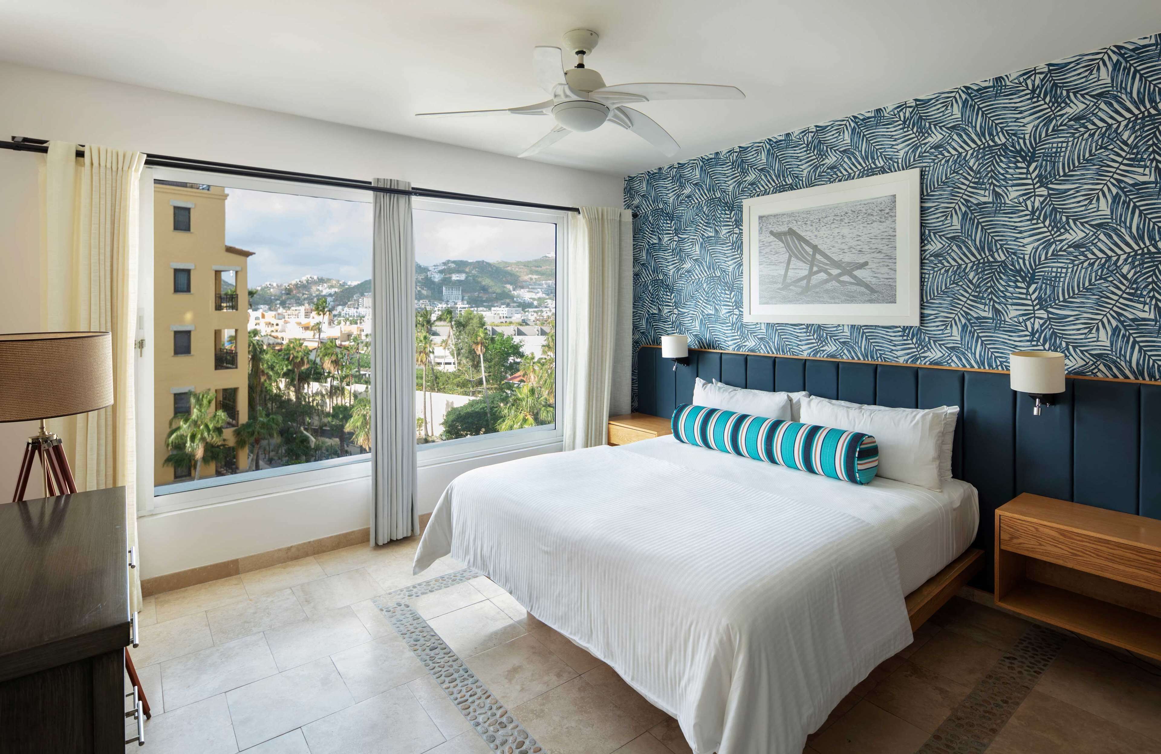 Ein Schlafzimmer mit Bett, Deckenventilator, großem Fenster mit Vorhängen und Blick auf die Stadt, sowie einer Wand mit Muster.