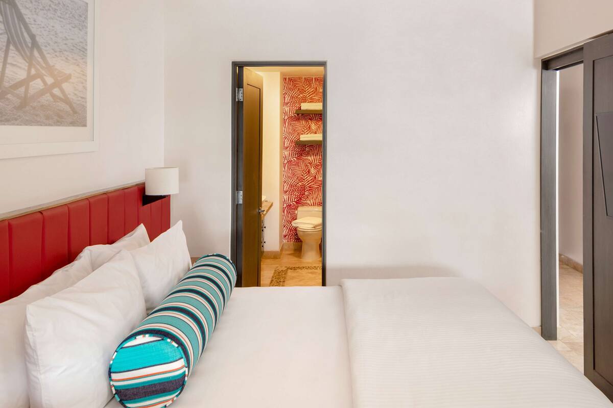 A hotel room with a bed, a red headboard, a striped pillow, a white lamp, and a view into the bathroom with a red and white patterned shower curtain.