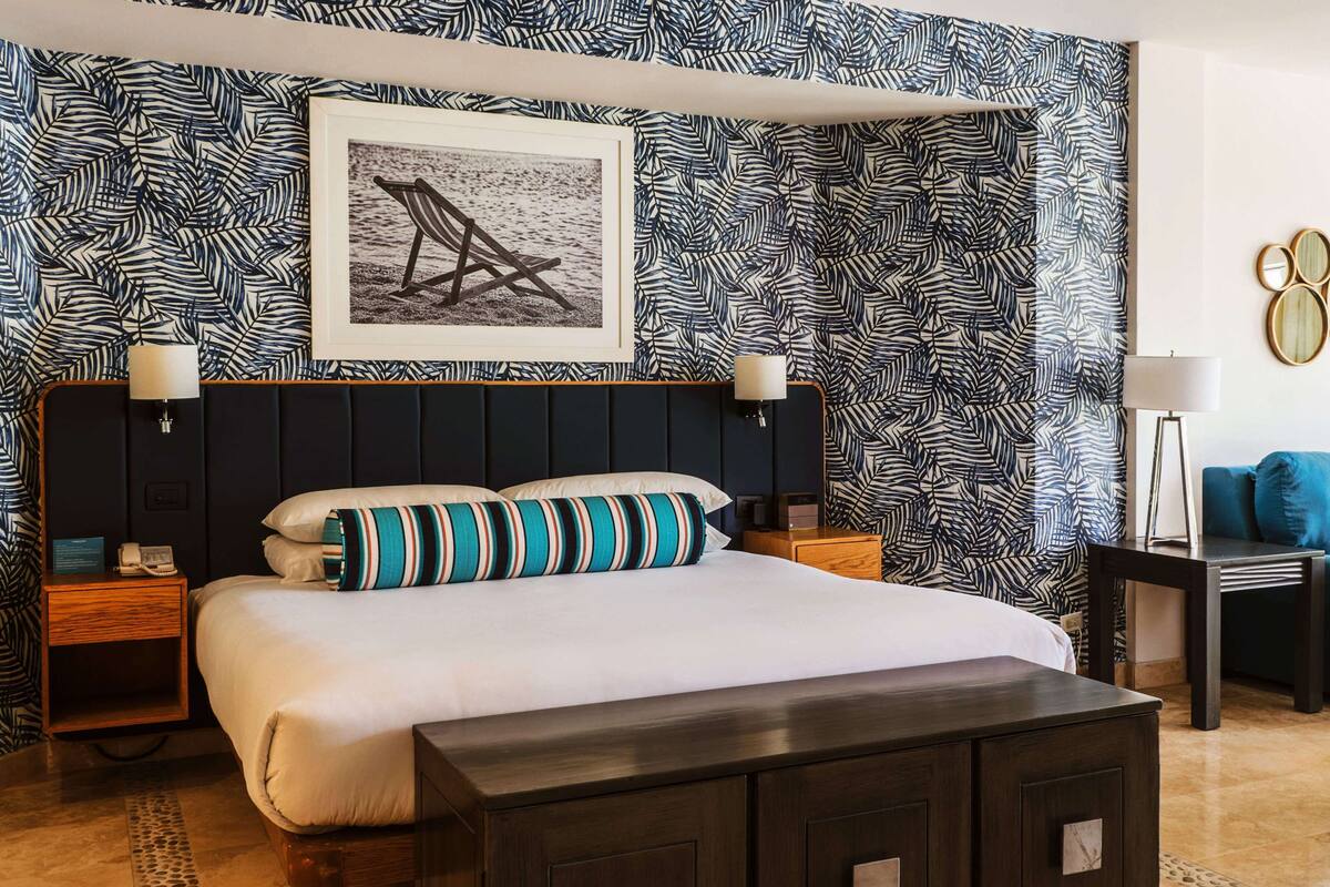 A hotel room with a bed, nightstands, a bench, a wall with a leaf pattern, a framed picture, and a blue armchair.