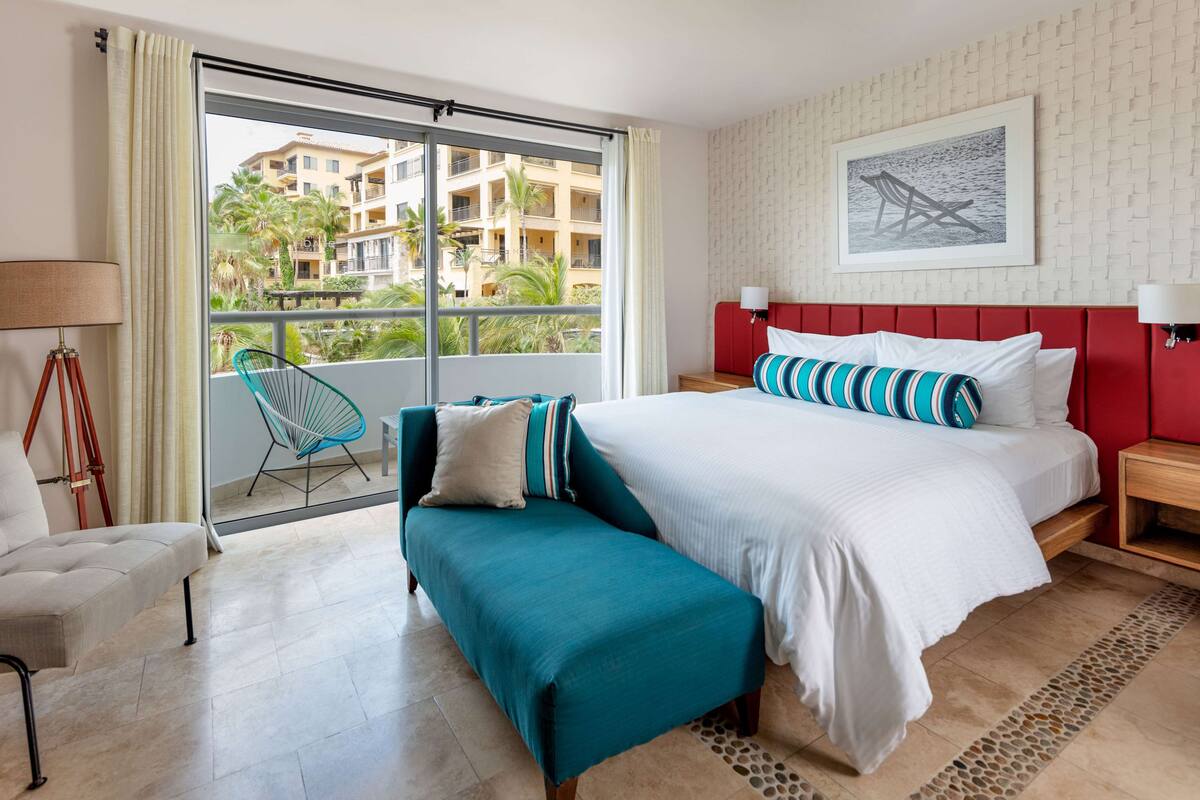 A hotel room with a large bed, a bench, a chair, and a view of a building with balconies.