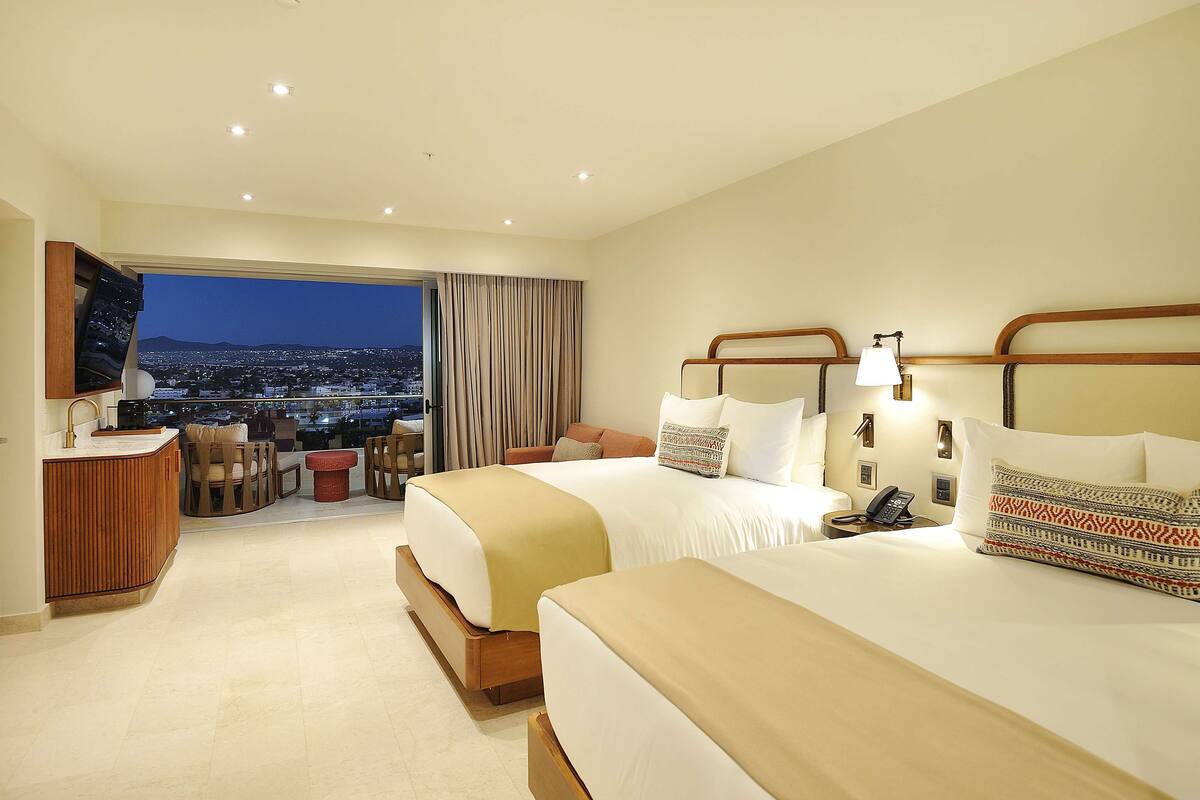 A hotel room with two beds, a TV, a desk, and a view of the city at night.