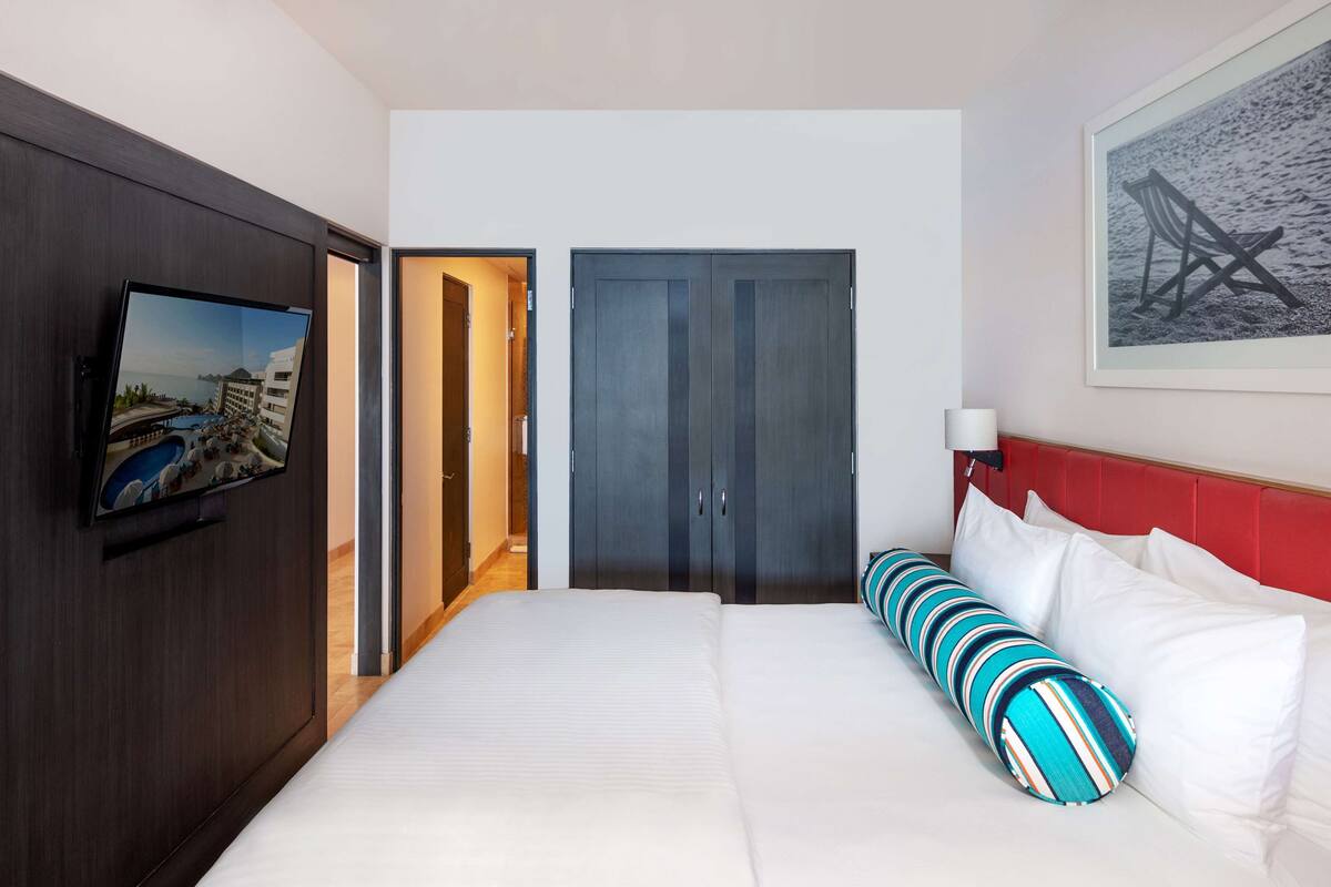 A hotel room with a bed, a TV mounted on the wall, a red headboard, and a framed picture on the wall.