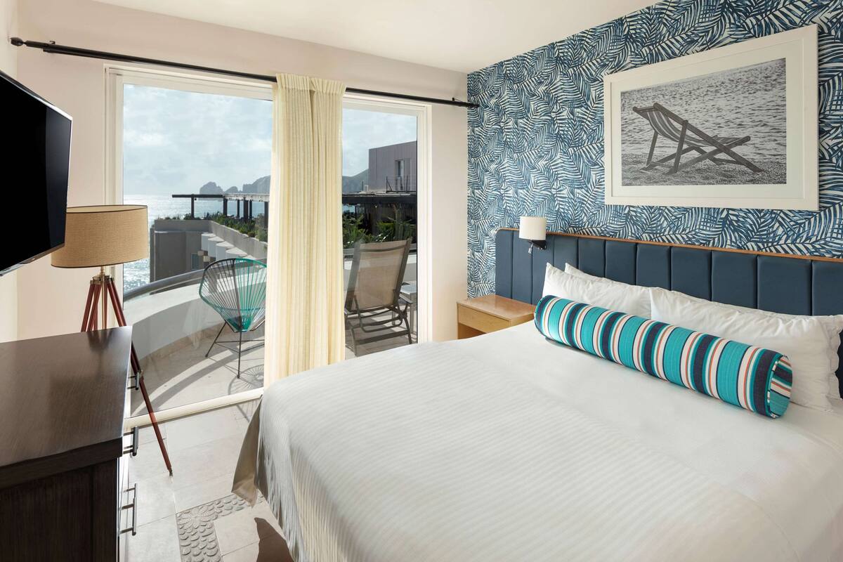 A hotel room with a large bed, a TV, a balcony with a chair, and a view of the sea.