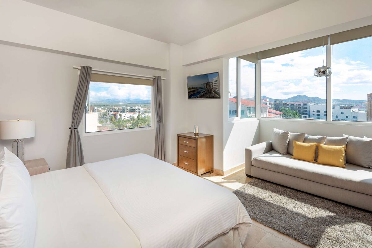 A hotel room with a large bed, a sofa, a nightstand, a lamp, a dresser, and a view of the city through large windows.