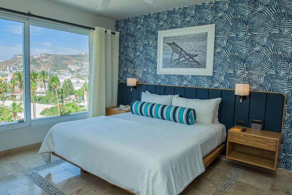 A bedroom with a large bed, two bedside tables, a nightstand, a window with a view of a cityscape, and a patterned wall.