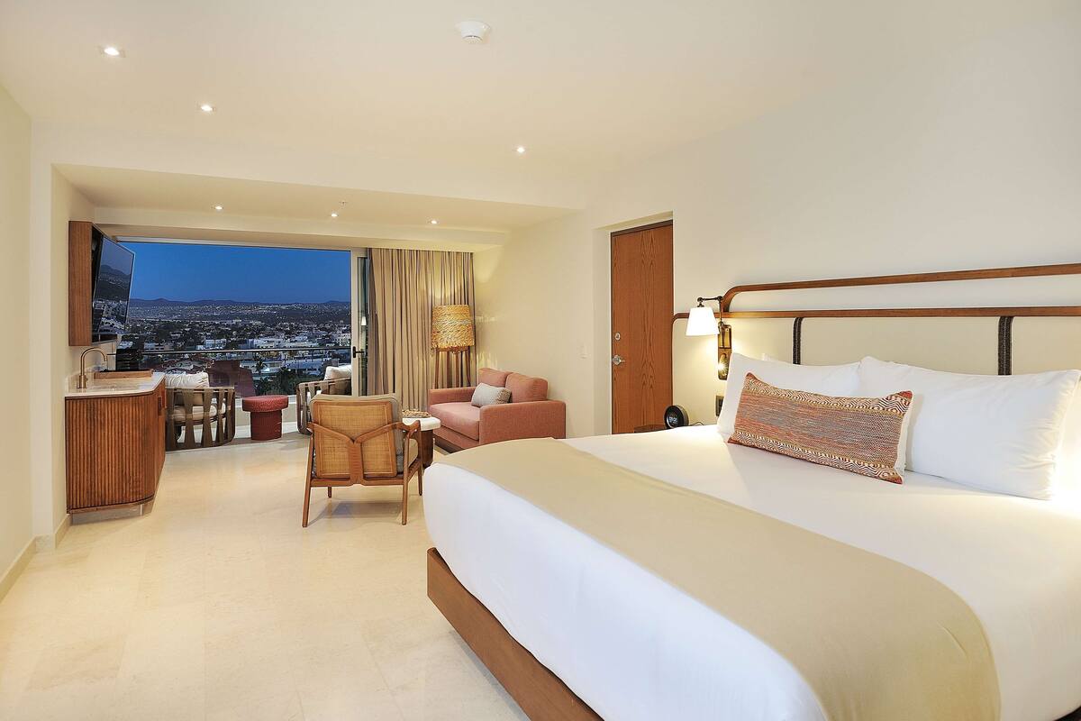 A hotel room with a large bed, a desk, a chair, and a view of a cityscape.