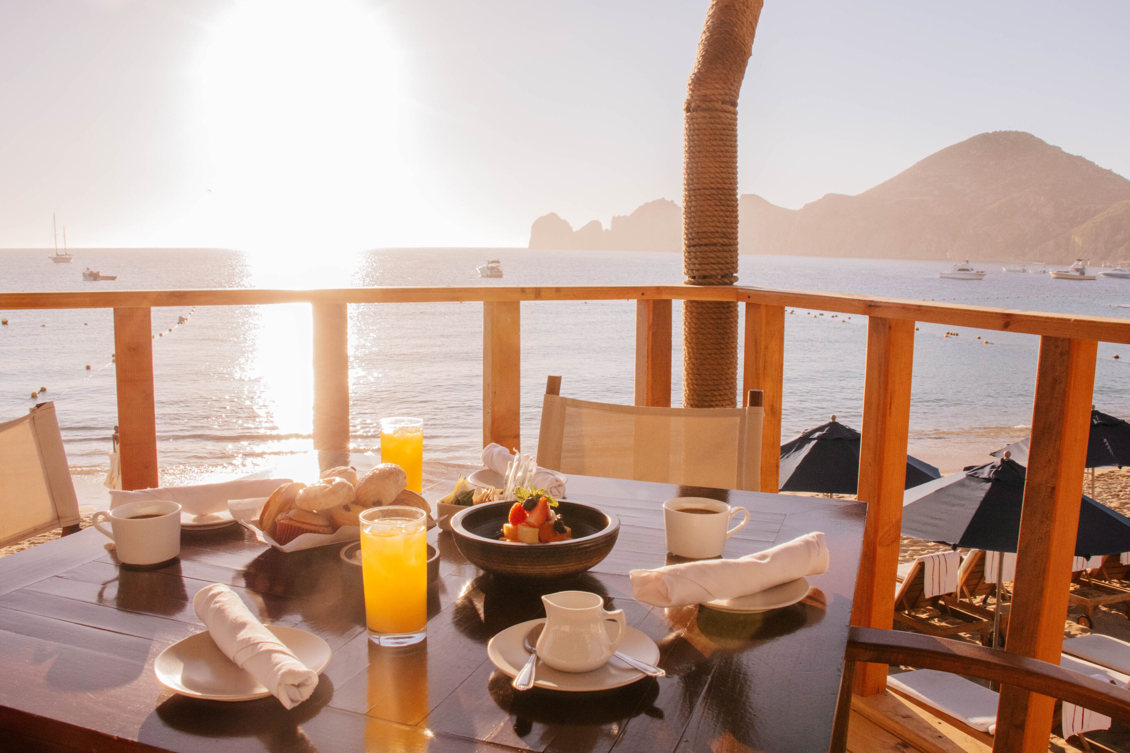 A dining table with breakfast items, a view of the sea, and beach umbrellas.