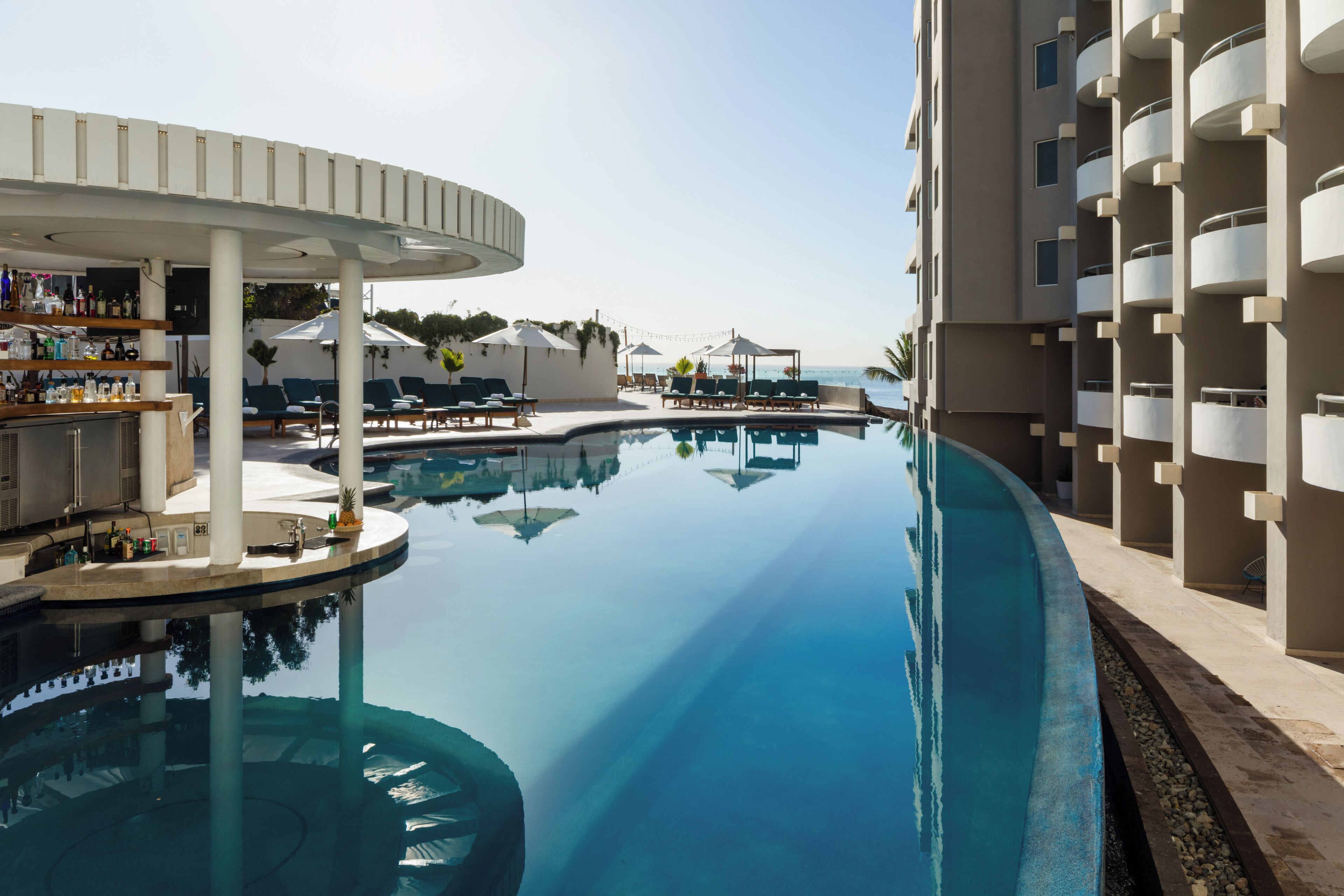 A pool area with lounge chairs and umbrellas, a bar with bottles, and a building with balconies.