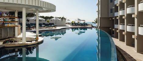 A pool area with lounge chairs and umbrellas, a bar with bottles, and a building with balconies.