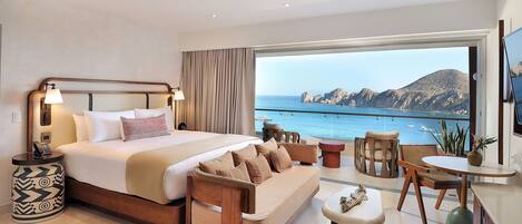 A modern hotel room with a large bed, a sofa, a dining area, and a view of the sea.