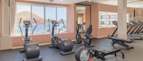 A gym with various cardio machines, large windows with a view of the sea, and a ceiling fan.