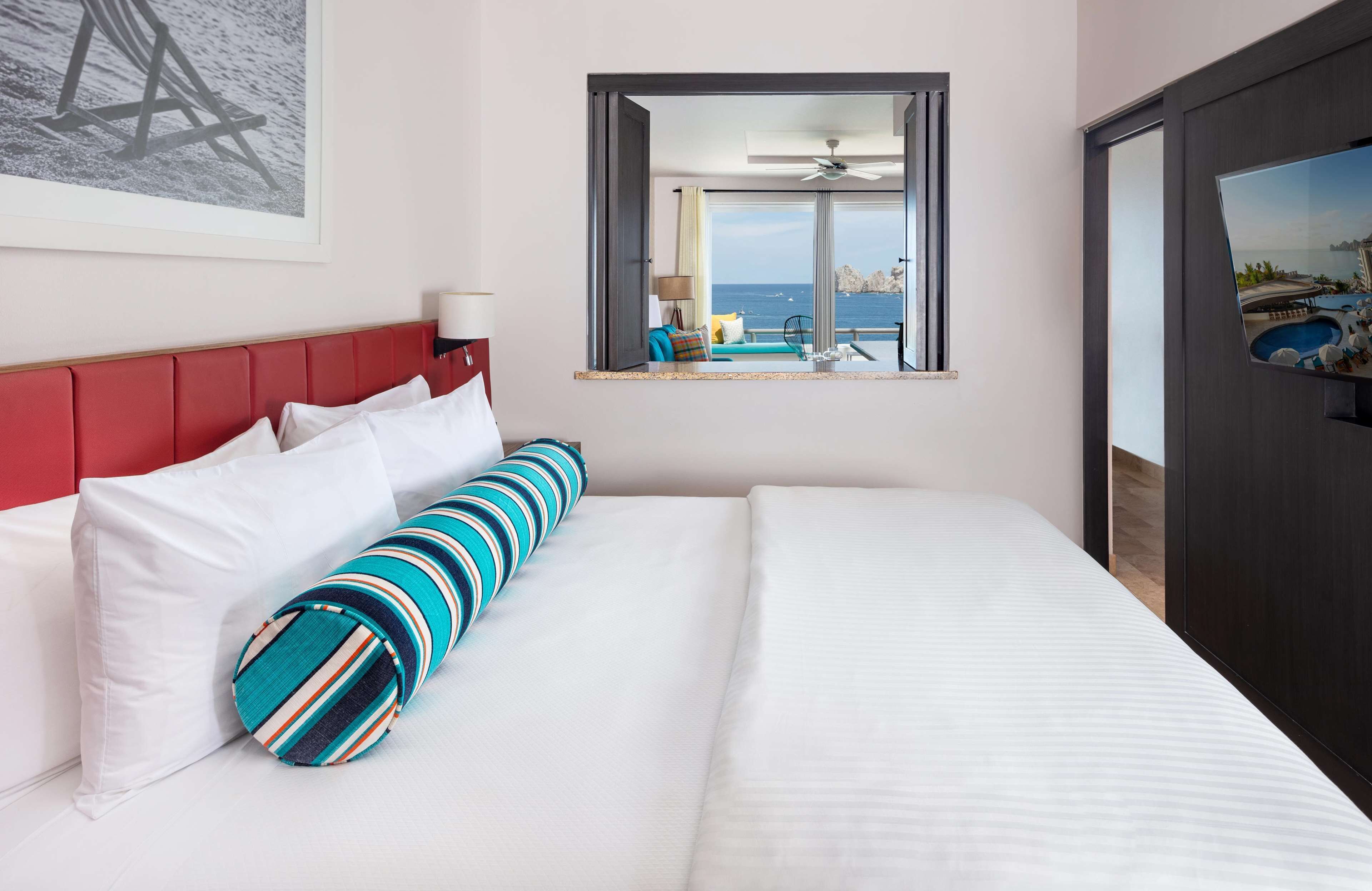 Une chambre d’hôtel avec un grand lit, une tête de lit rouge et une vue sur la mer par la fenêtre.