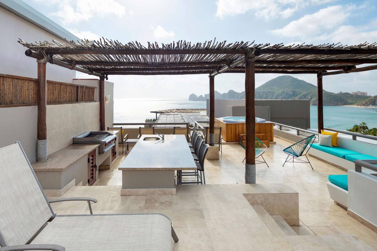 An outdoor patio with a long table, chairs, a barbecue grill, a hot tub, and a view of the sea.