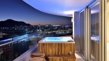 A modern balcony with a hot tub, wooden stools, and a city view at dusk.