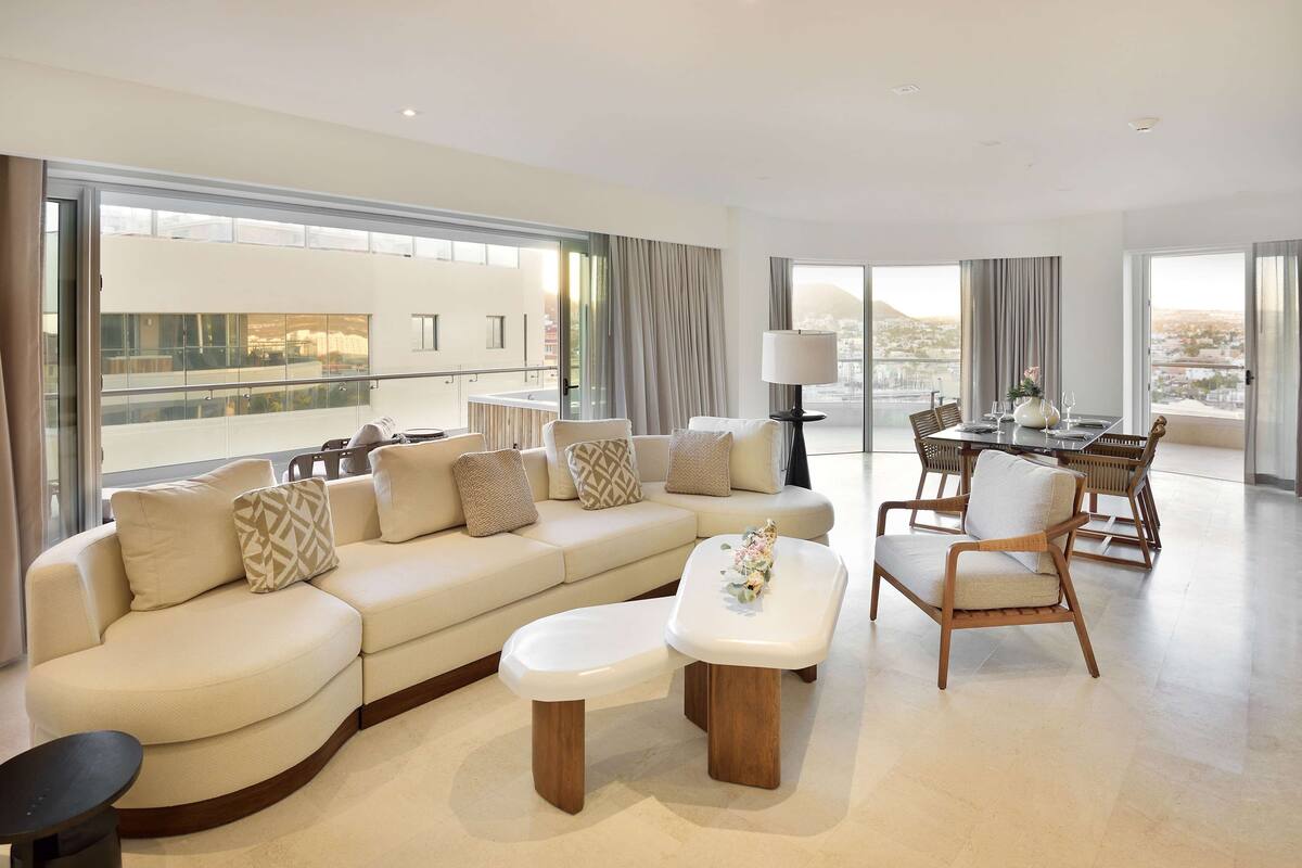 A modern living room with a sofa, ottoman, and chairs, large windows, and a dining area.
