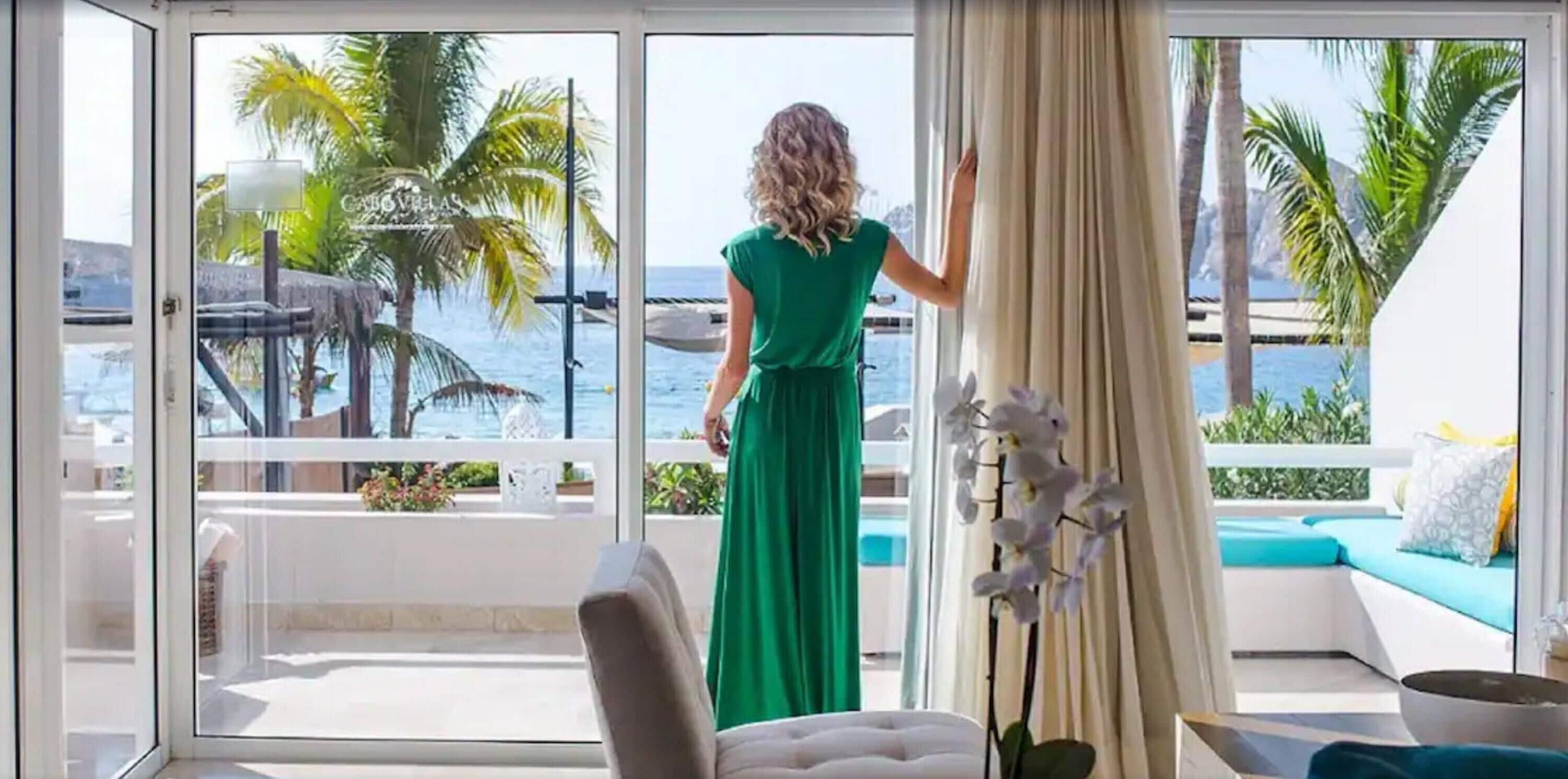 A room with a large sliding glass door leading to a balcony with a view of the sea and palm trees.