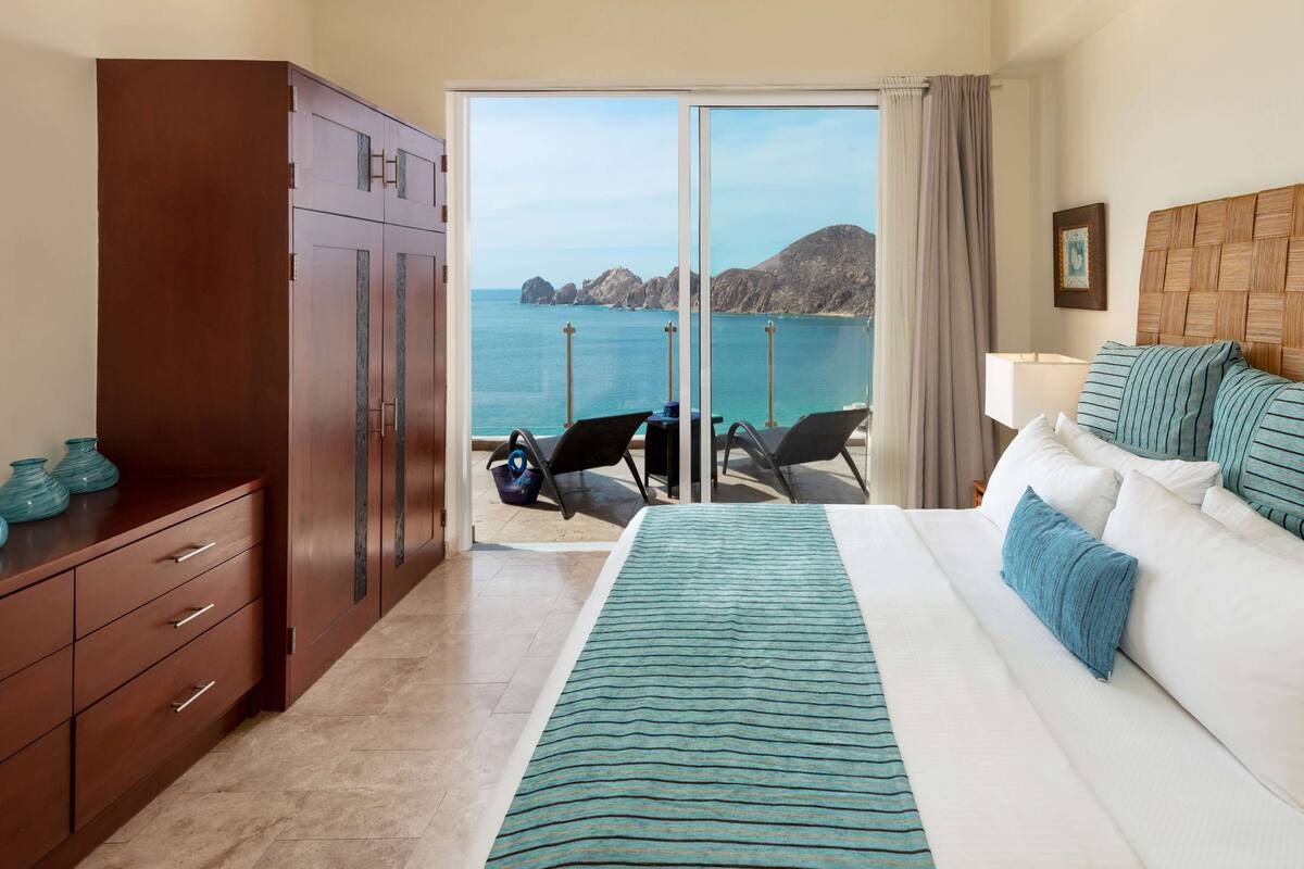 A hotel room with a bed, a wooden wardrobe, a desk with chairs, and a view of the sea.
