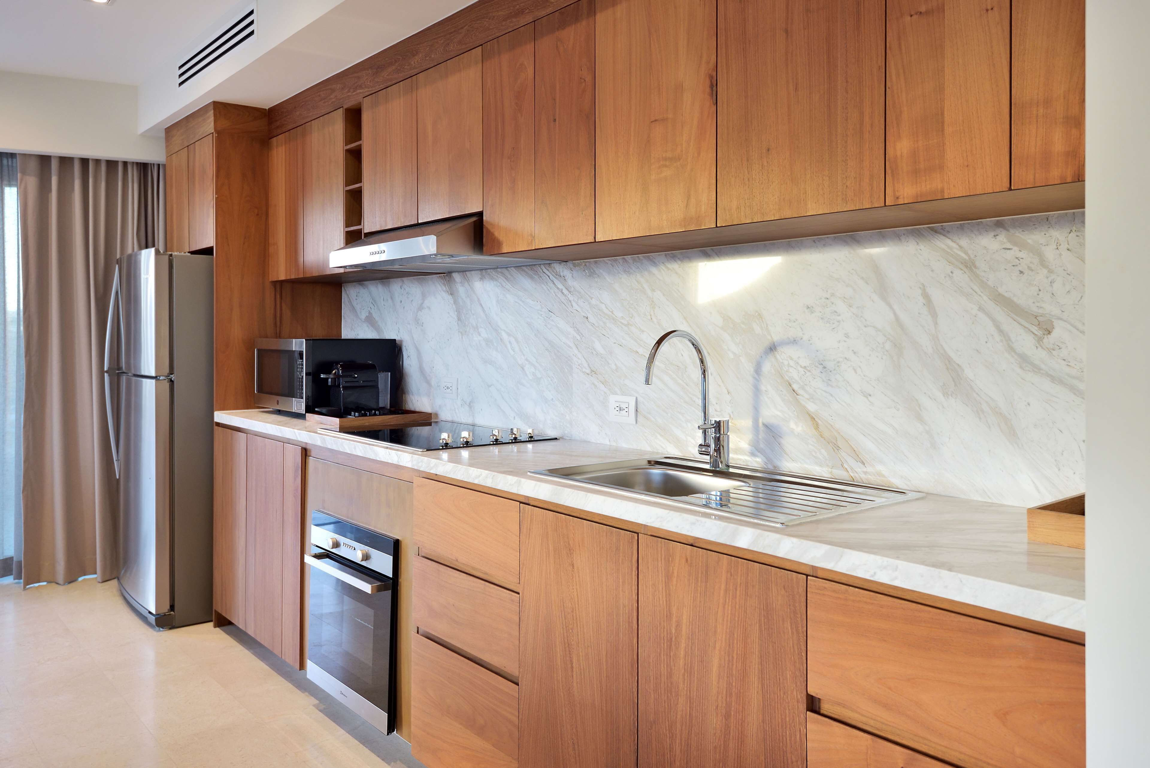 A modern kitchen with wooden cabinets, a stainless steel refrigerator, a microwave, and a sink with a marble countertop.