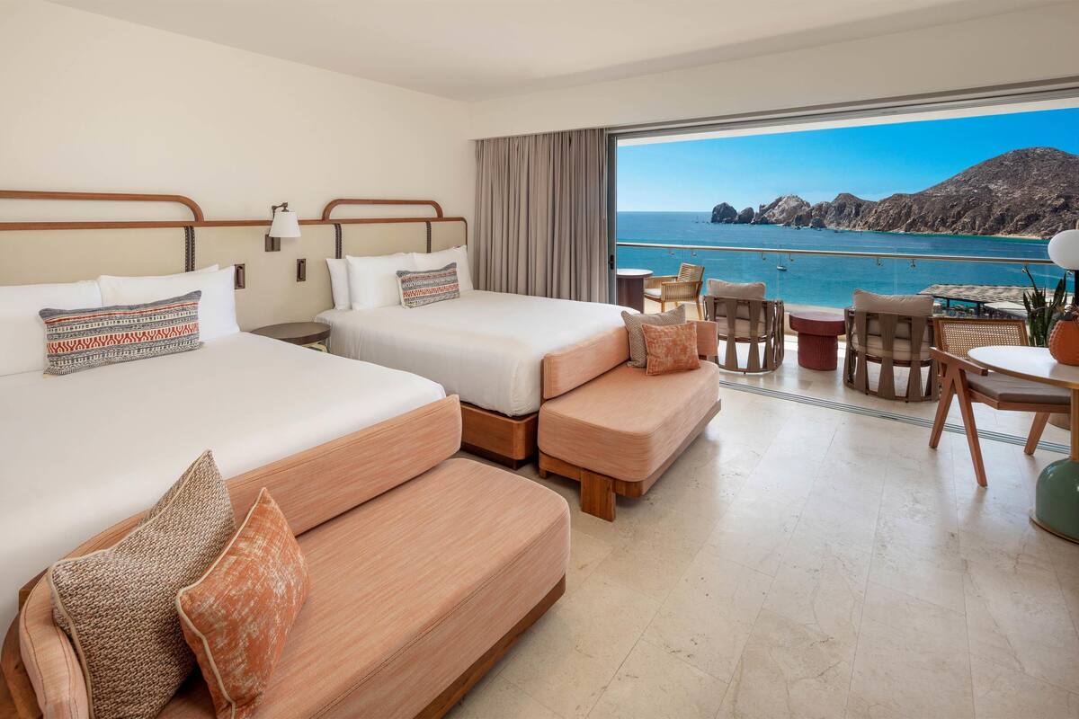 A hotel room with two beds, a balcony with a view of the sea, and a dining area with wicker furniture.