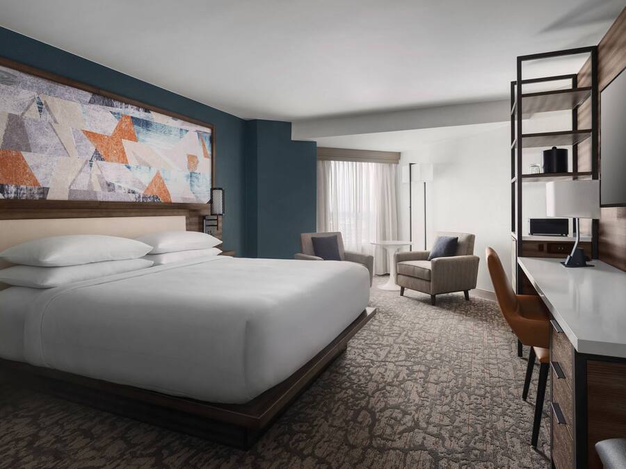 A hotel room with a large bed, a desk, two chairs, a small table, and a large abstract painting on the wall.