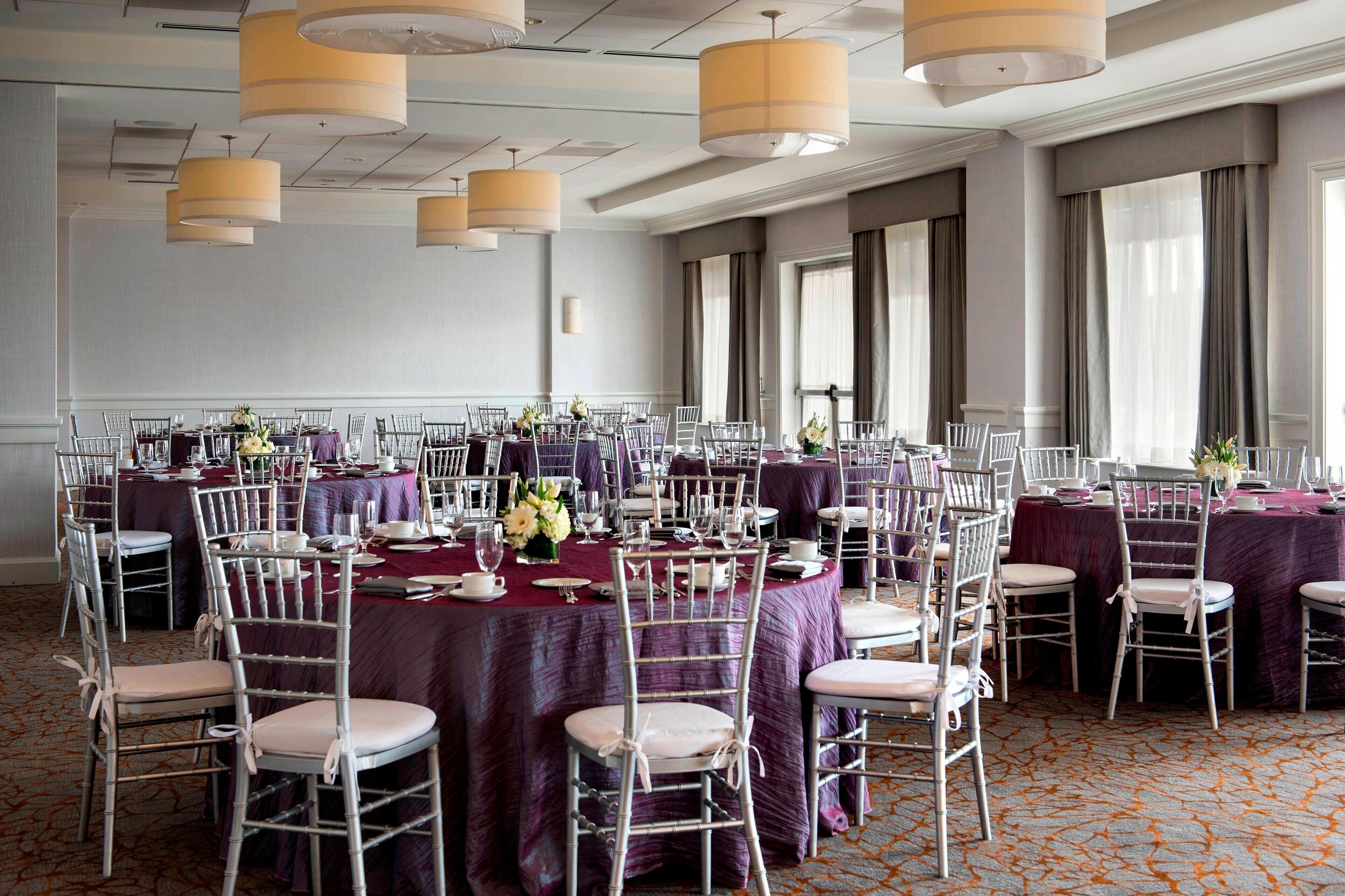 Une salle de banquet avec des tables rondes recouvertes de nappes violettes, des chaises blanches et des suspensions lumineuses jaunes.