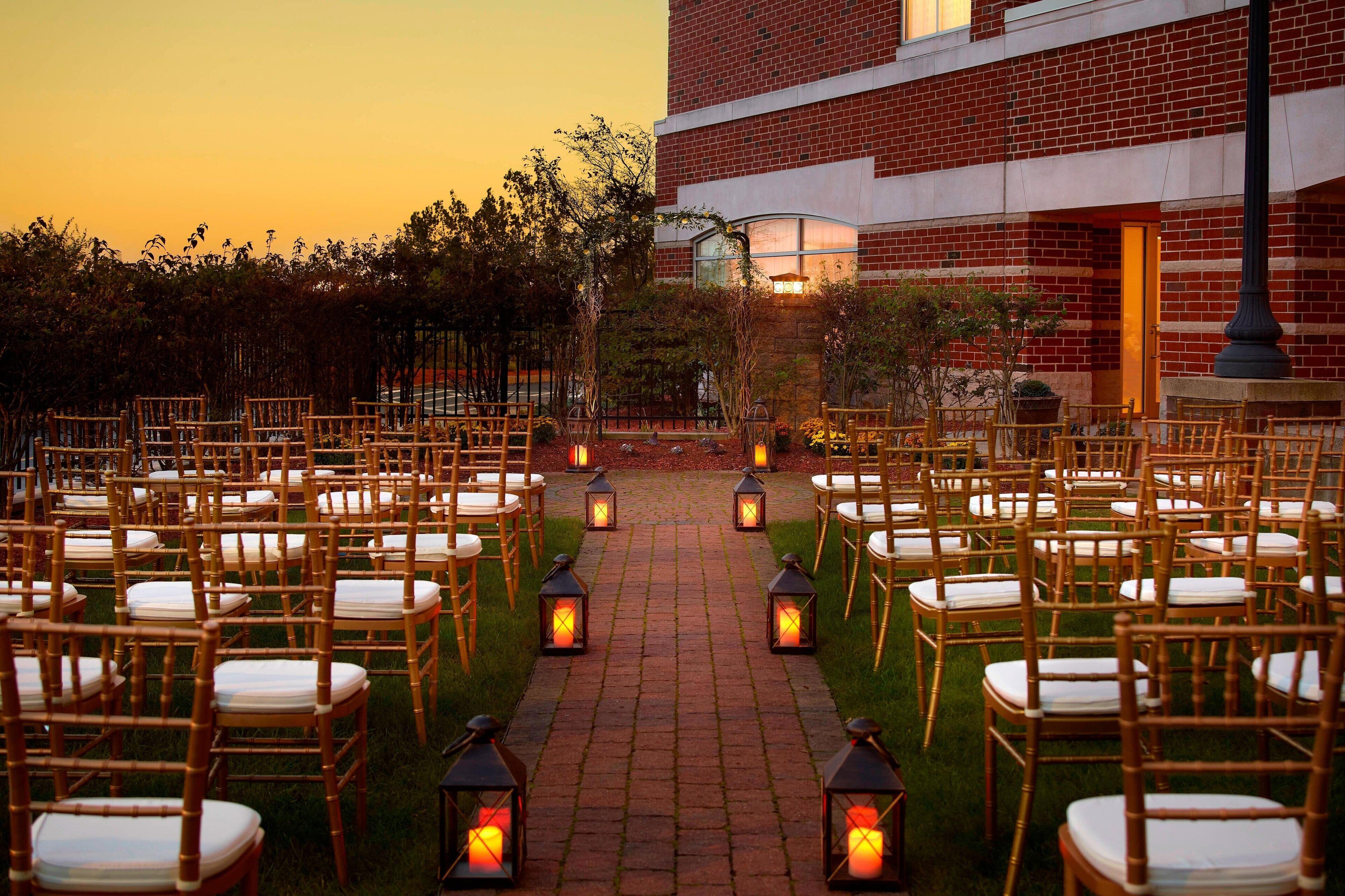 A brick building with a paved pathway lined with chairs and lanterns, suggesting an outdoor event or ceremony area.