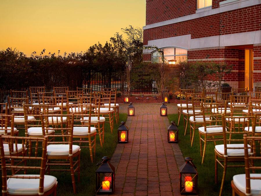 Een bakstenen gebouw met een verhard pad bezaaid met stoelen en lantaarns, wat wijst op een evenementen- of ceremonieterrein buiten.