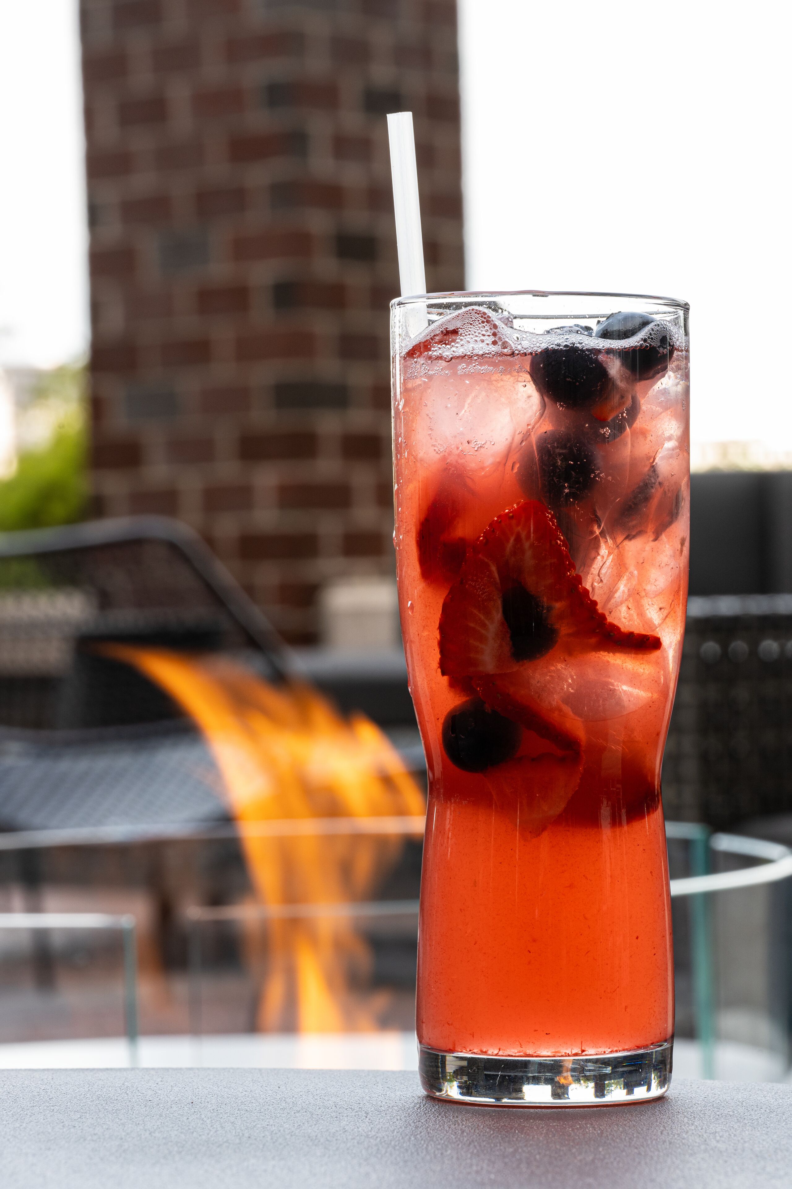 A tall glass with a straw, filled with a red beverage and ice, placed on a table with a blurred background of a brick building and a car.