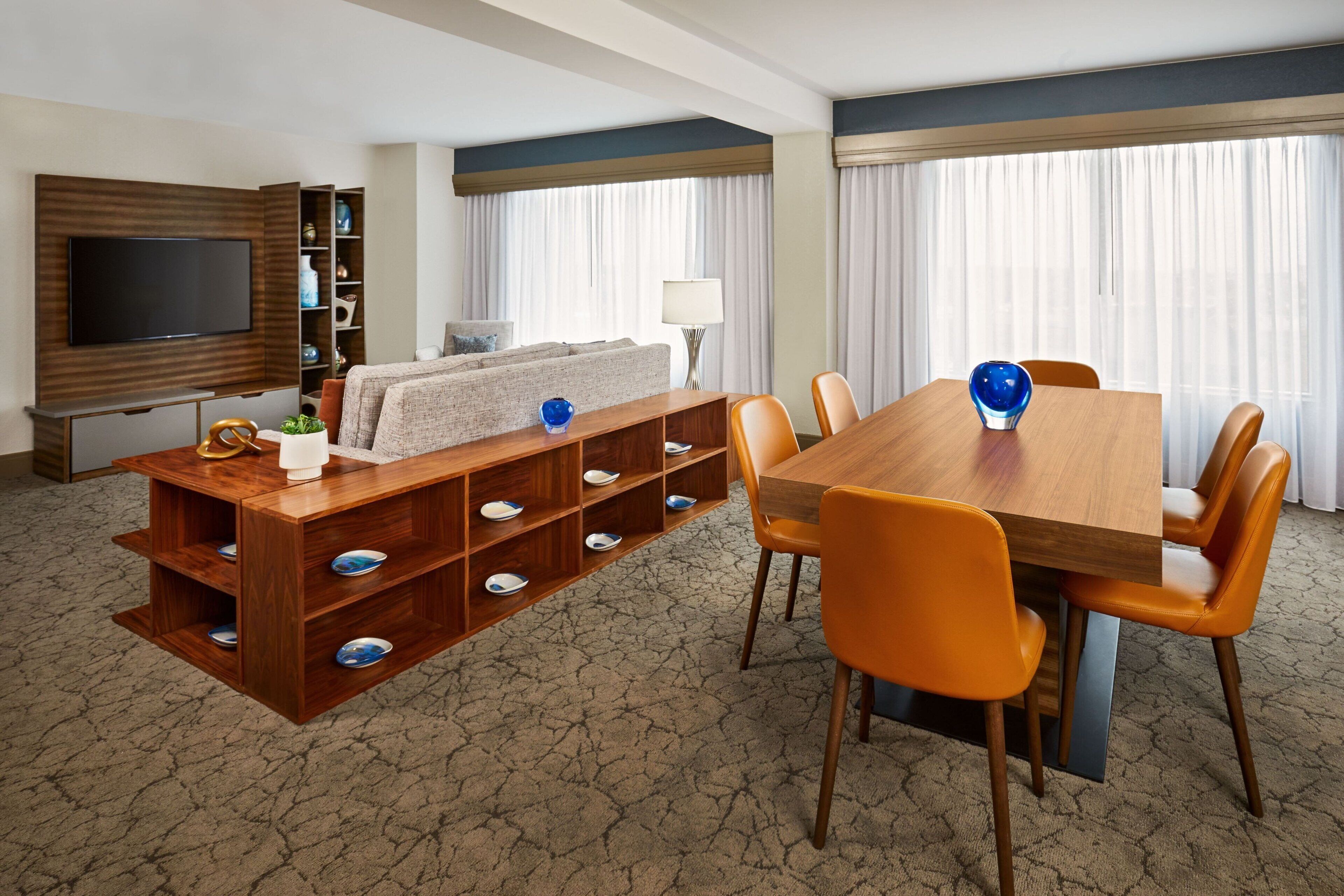 A hotel room with a wooden dining table, orange chairs, a TV mounted on a wooden panel, a sofa, a bookshelf, and a large window with curtains.