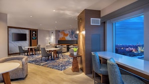 Un hall d’hôtel moderne doté d’une grande fenêtre offrant une vue sur la ville, d’un téléviseur à écran plat fixé au mur et de différents types de sièges, notamment des canapés et des fauteuils.