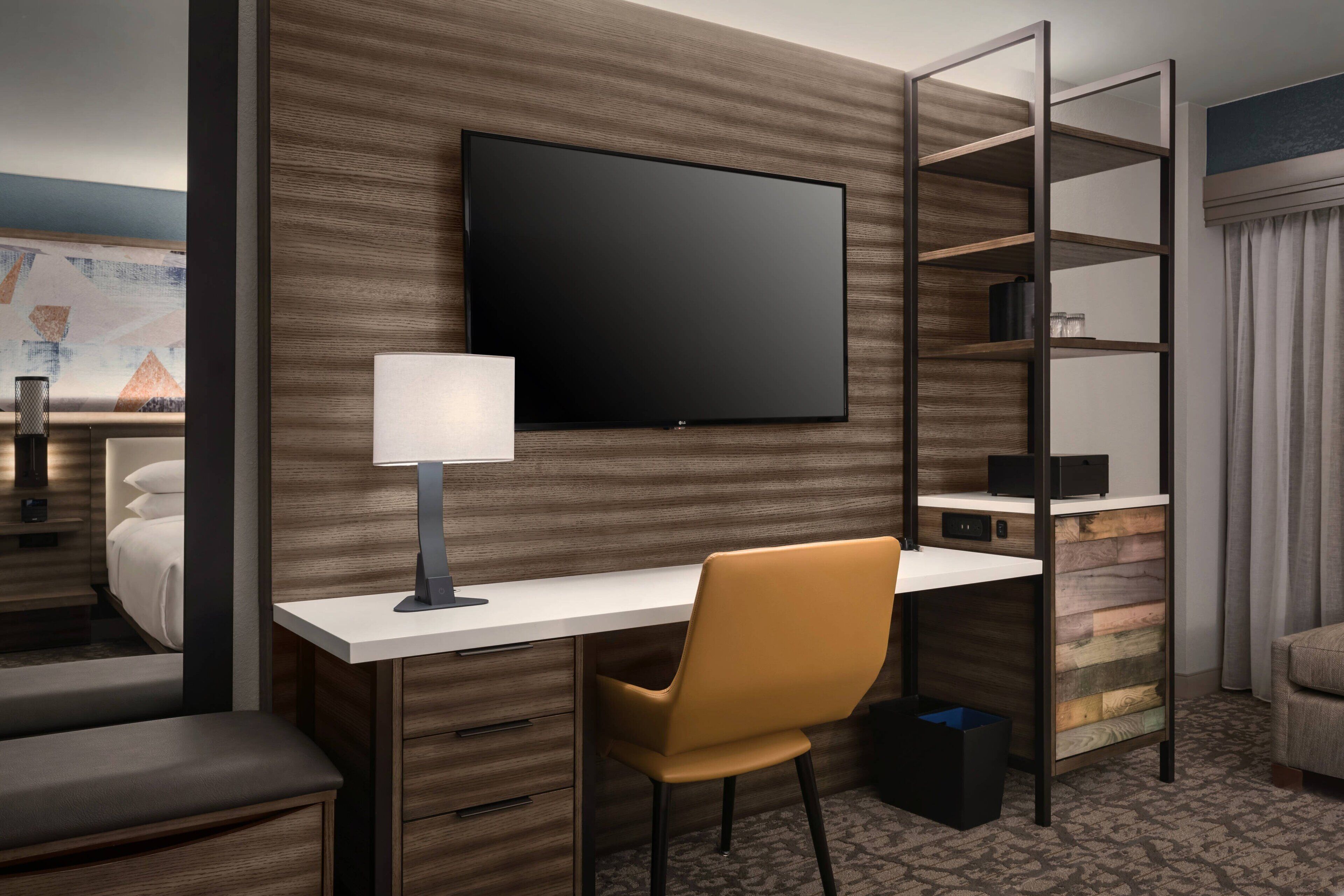 A hotel room with a flat-screen TV mounted on a wooden wall, a desk with a lamp, a chair, and a built-in bookshelf.