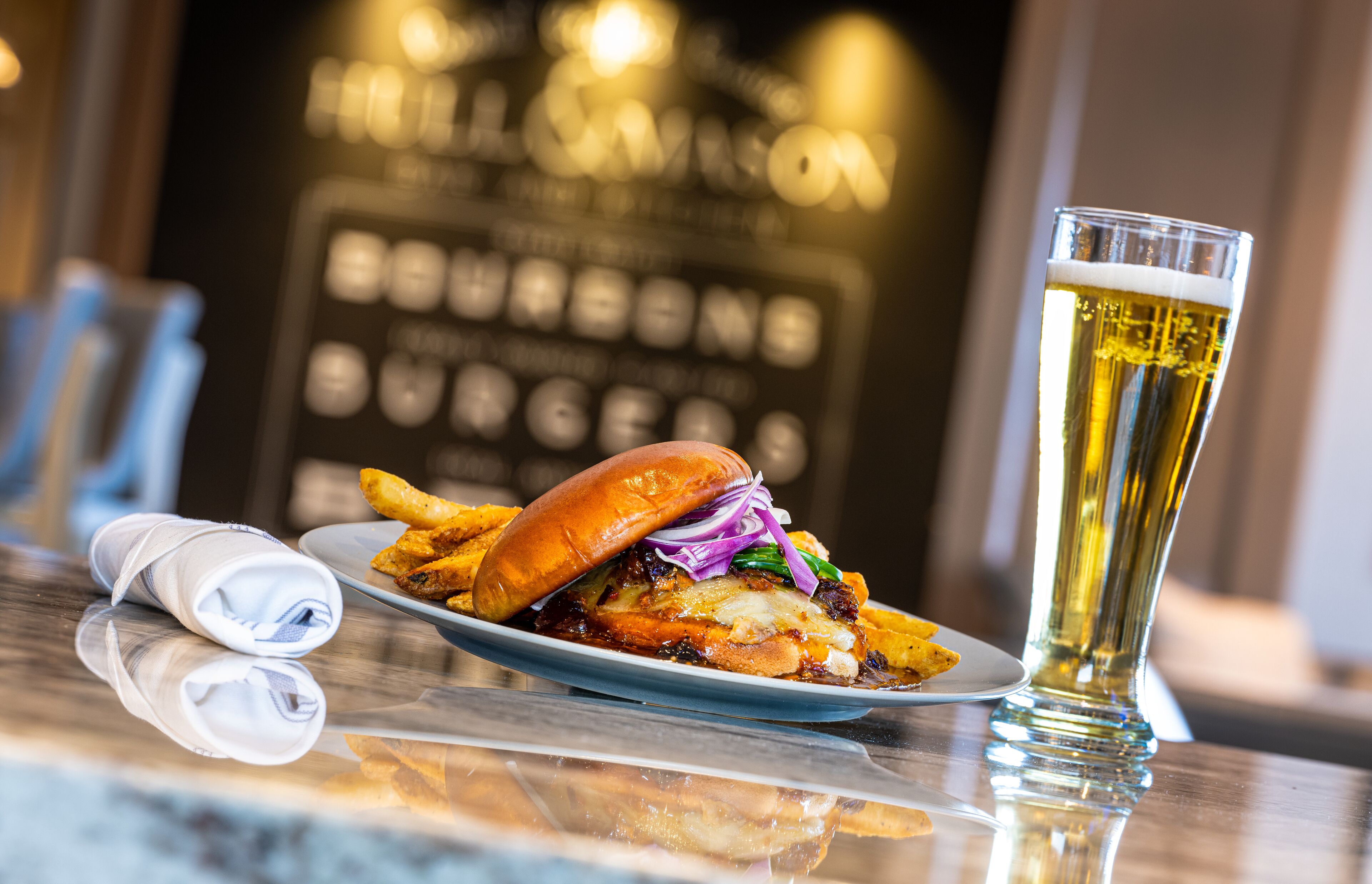 Um prato com hambúrguer e batatas fritas, um copo de cerveja e um guardanapo sobre a mesa.