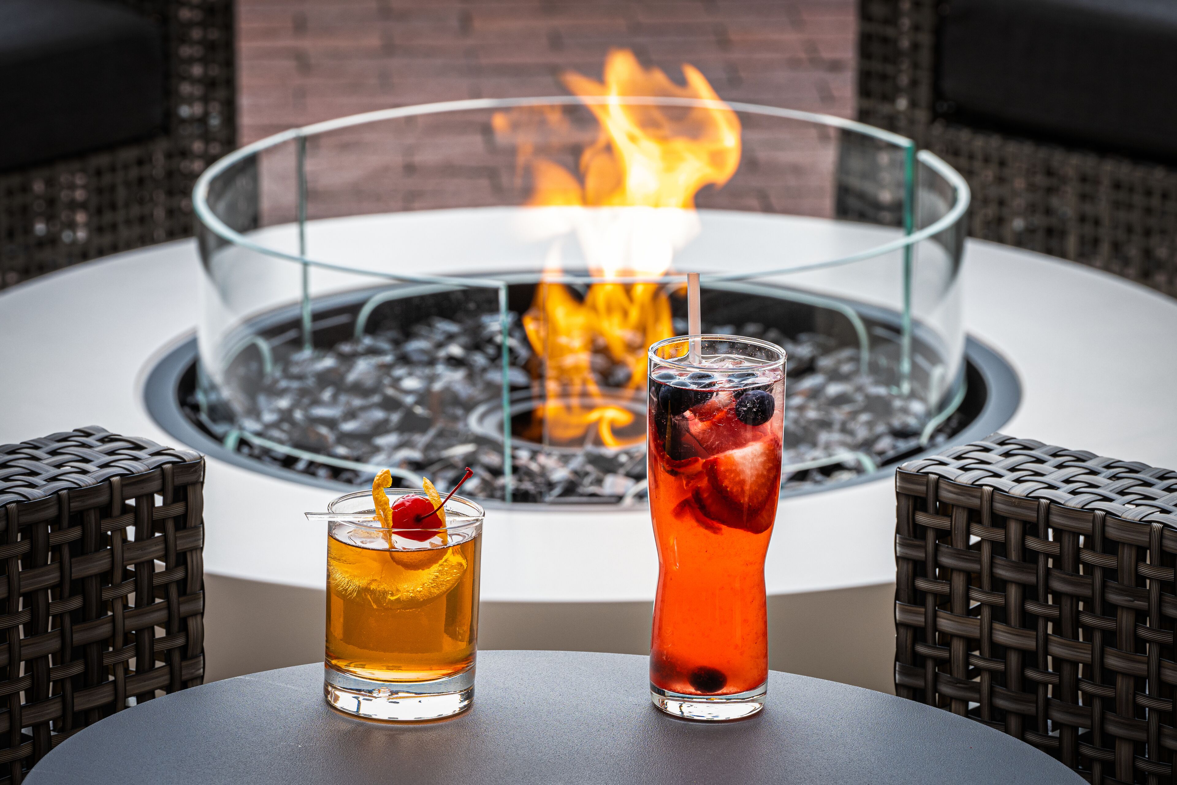 Two drinks on a table with a glass fire pit in the background.