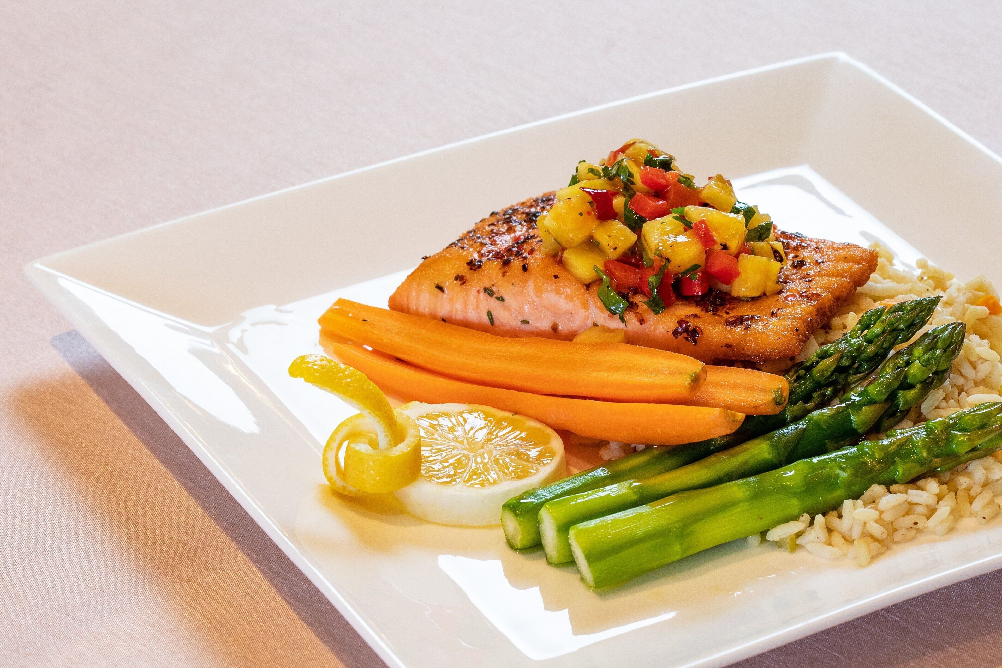Un piatto bianco con un filetto di pesce alla griglia, condito con salsa di mango, asparagi e fette di limone.