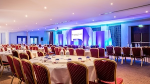 Un salón de conferencias con filas de mesas y sillas, un escenario con una pantalla, y un cartel para Crowne Plaza Hotel & Resorts.