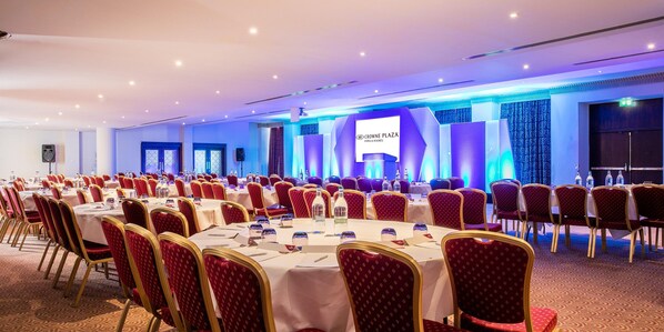 A conference room with rows of tables and chairs, a stage with a screen, and a sign for Crowne Plaza Hotel & Resorts.