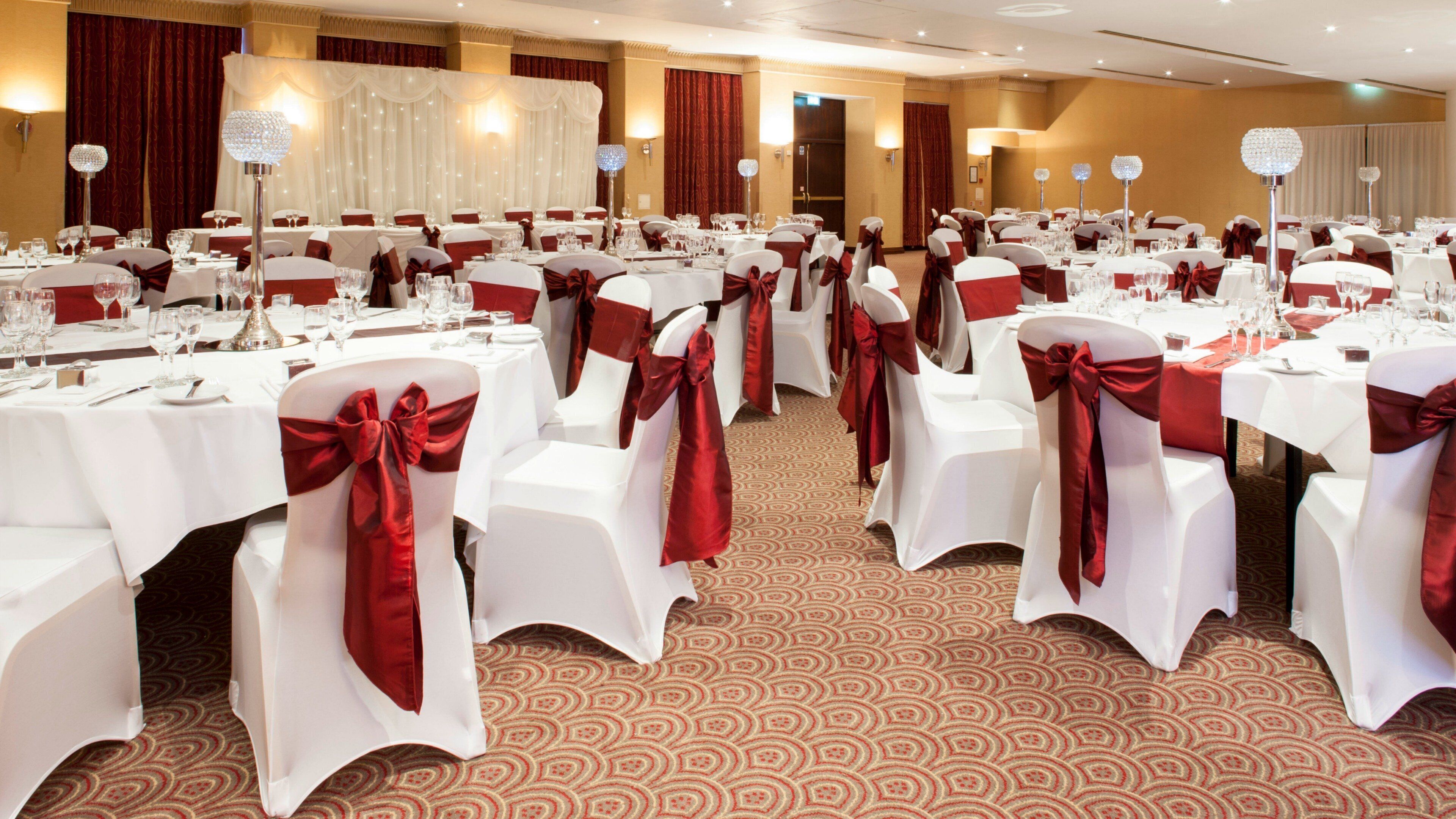 Un salón de banquetes con mesas redondas, manteles blancos y sillas con cintas rojas.