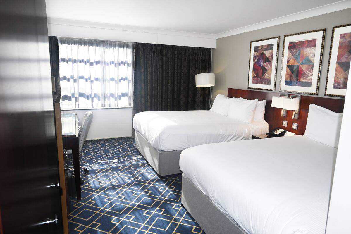 A hotel room with two beds, a desk, a chair, and a window with curtains. There are three framed artworks on the wall.