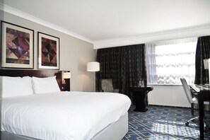 Ein Hotelzimmer mit einem großen Bett, zwei gerahmten Bildern an der Wand, einem Schreibtisch mit Stuhl, einer Lampe und einem Fenster mit Vorhängen.