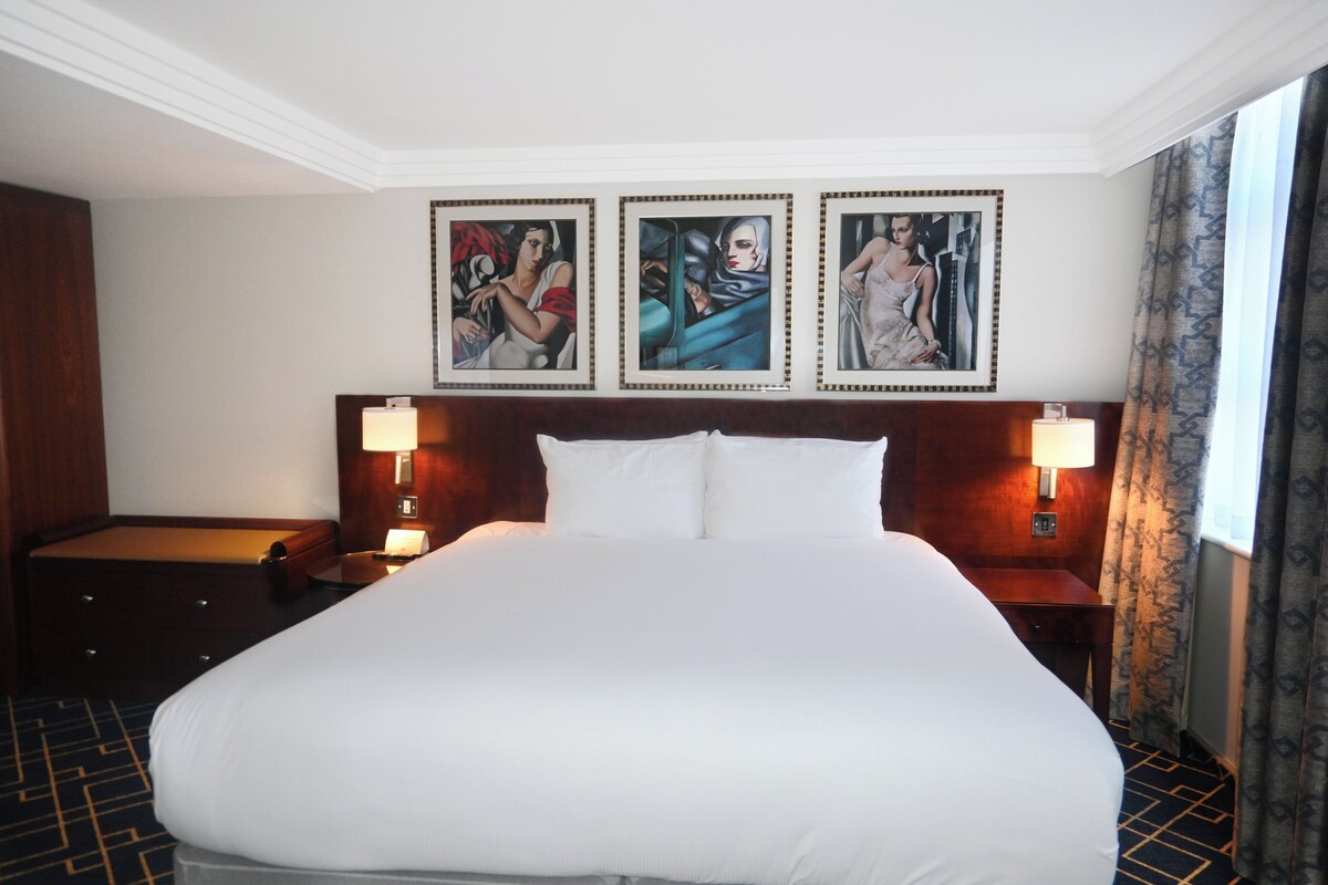 Une chambre d’hôtel avec un grand lit, deux tables de chevet avec des lampes, une tête de lit en bois et trois œuvres d’art encadrées accrochées au mur.