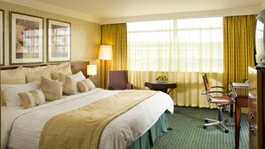 A hotel room with a large bed, a desk with a chair, a small table with a fruit bowl, and a television.